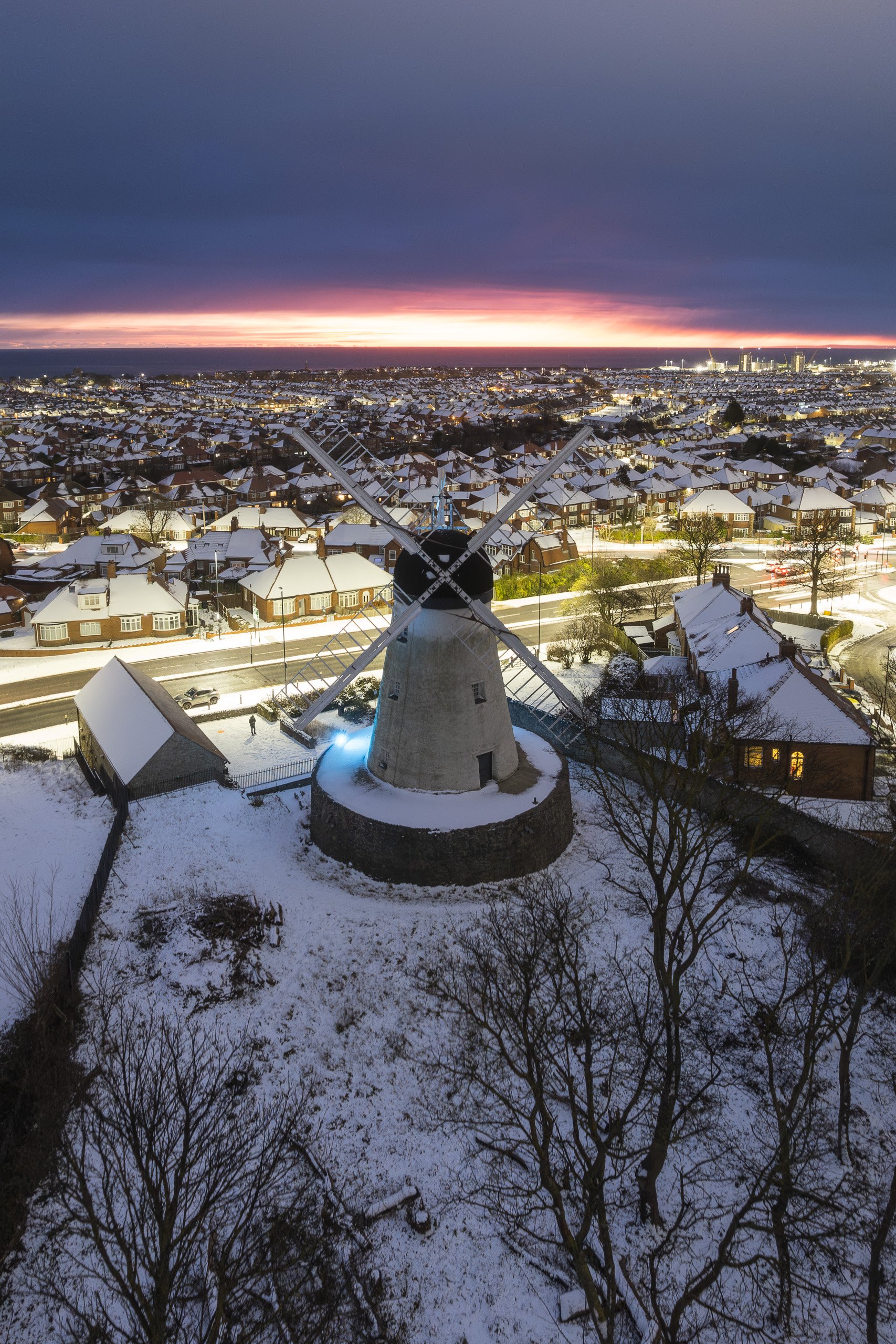 Aerial view of Fulwell Mill windmill standing over snow-covered rooftops at dusk, a vivid pink sunset glowing on the horizon.