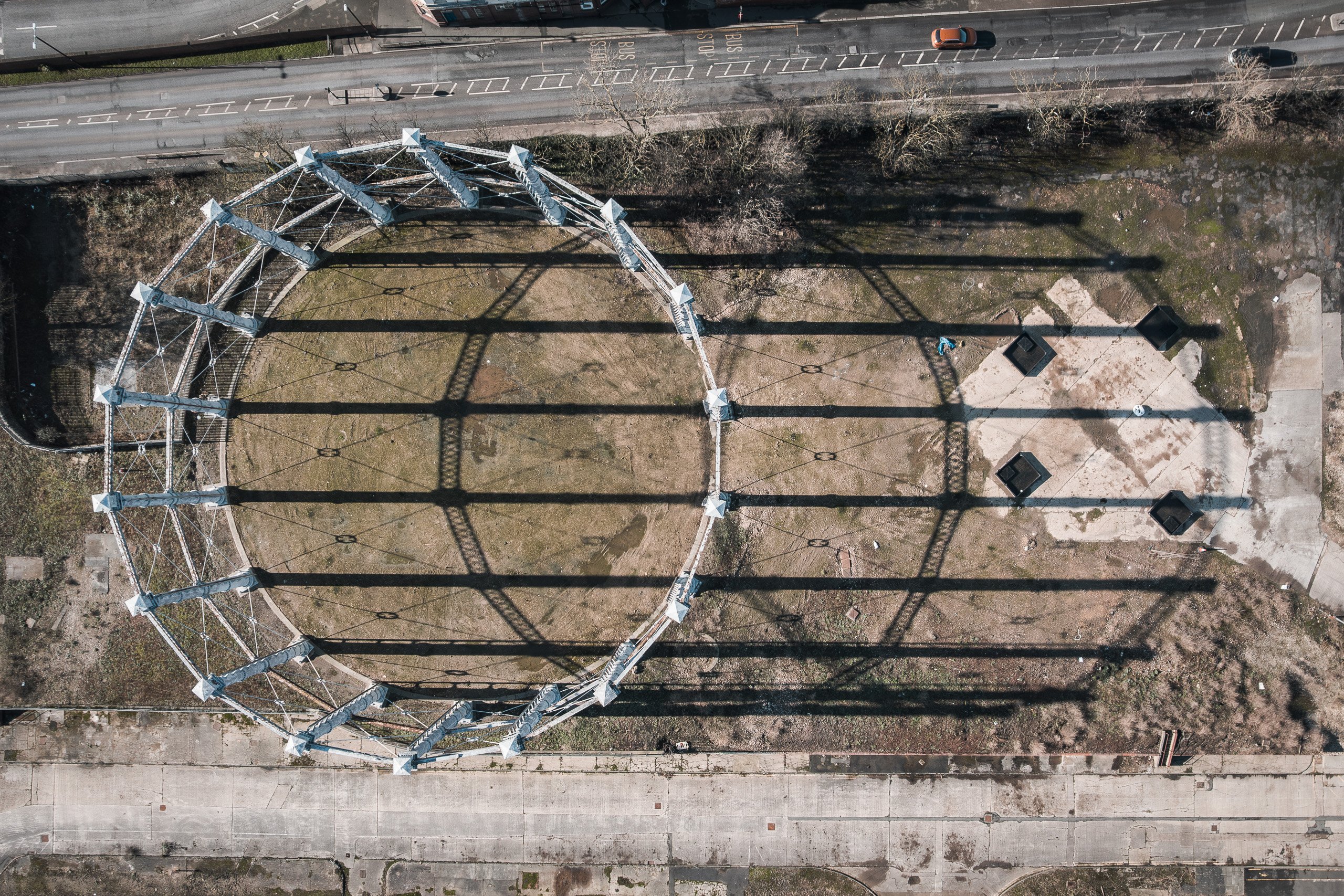 Top-down aerial view of the skeletal frame of a disused gas holder, casting long geometric shadows across cleared ground.