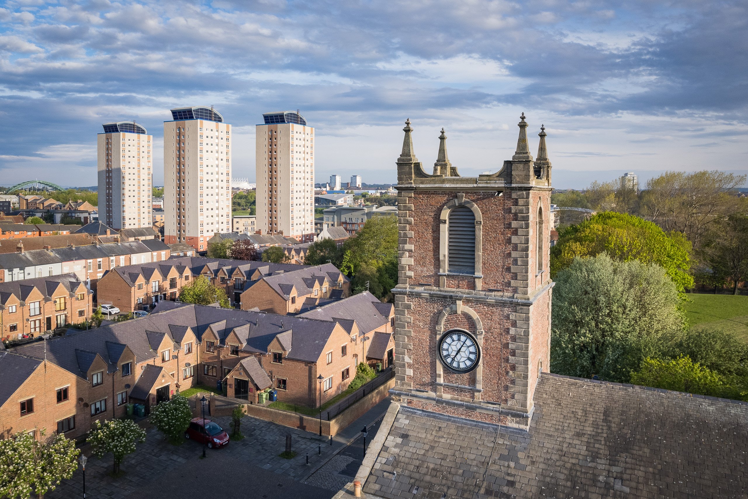 Aerial close-up of the clock tower of the old Holy Trinity Church, its red-brick and stone pinnacles rising above the East End, high-rise residential towers in the background.