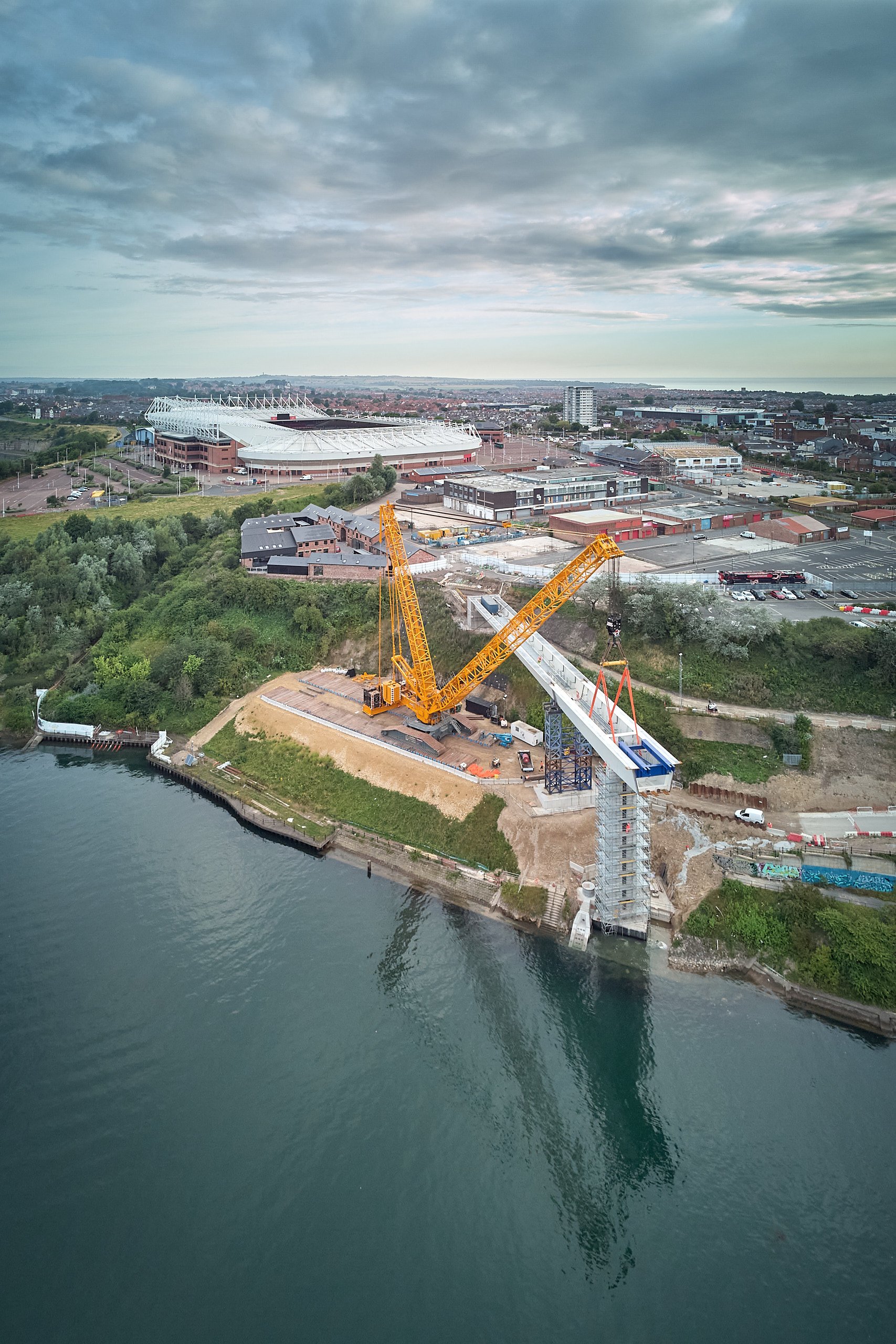 Aerial view of a crane lifting a bridge section into place during construction of the Keel Crossing over the River Wear, a football stadium visible in the background.
