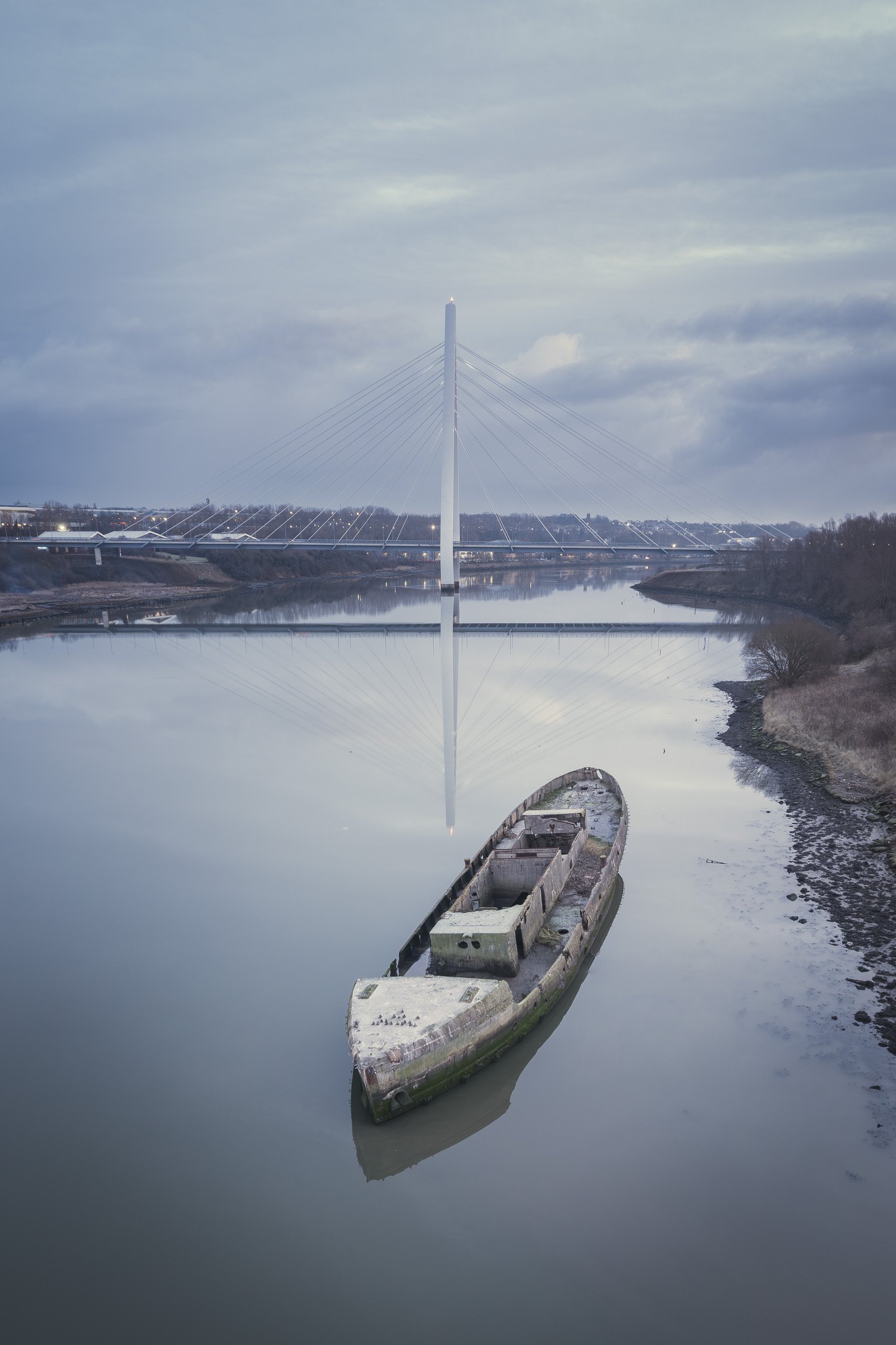 Aerial view of a derelict boat hull resting in still river water, the Northern Spire Bridge and its reflection rising behind at dusk.