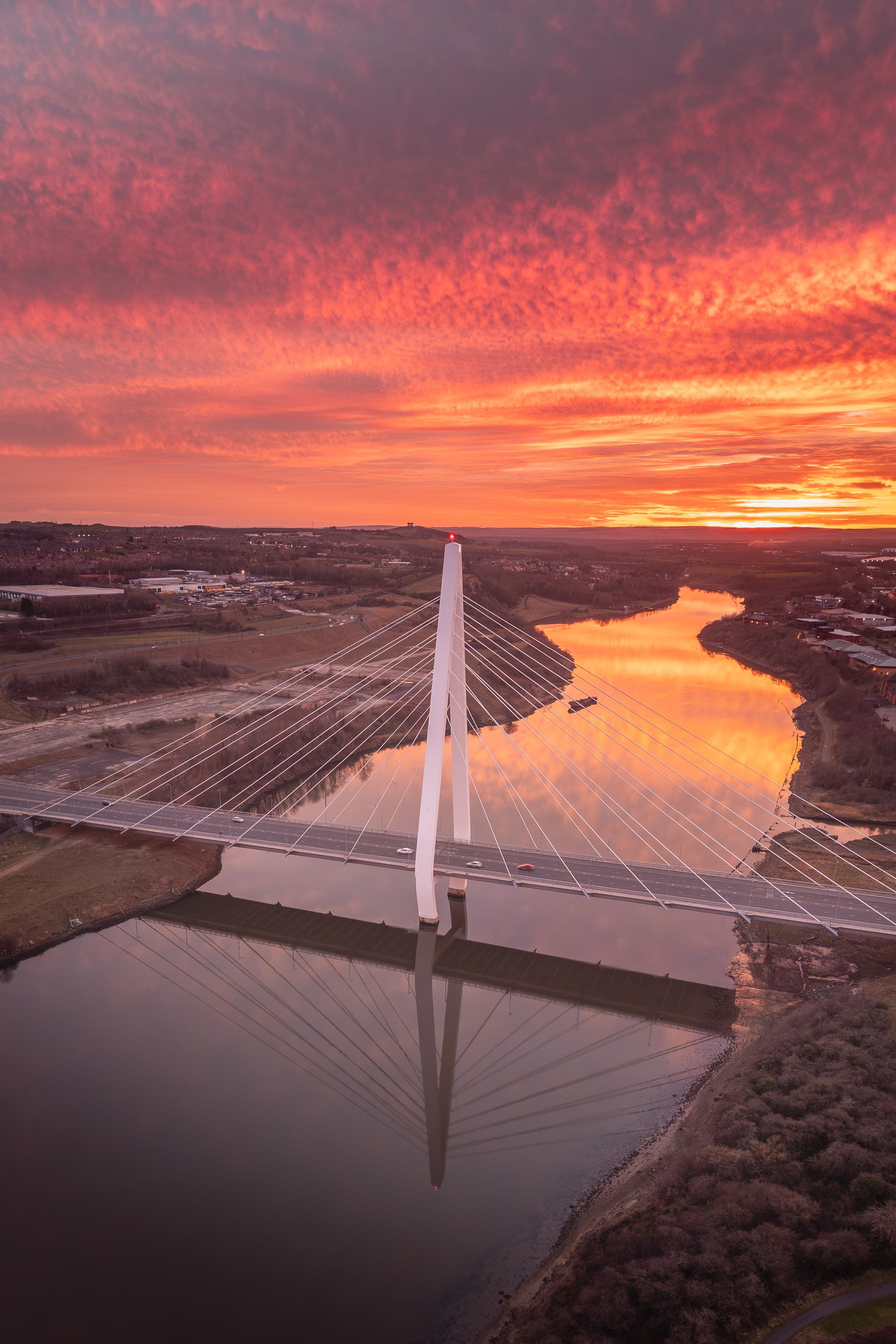 Aerial view of the Northern Spire Bridge spanning the River Wear beneath a vivid red and orange sunset sky, its cables mirrored in the water.