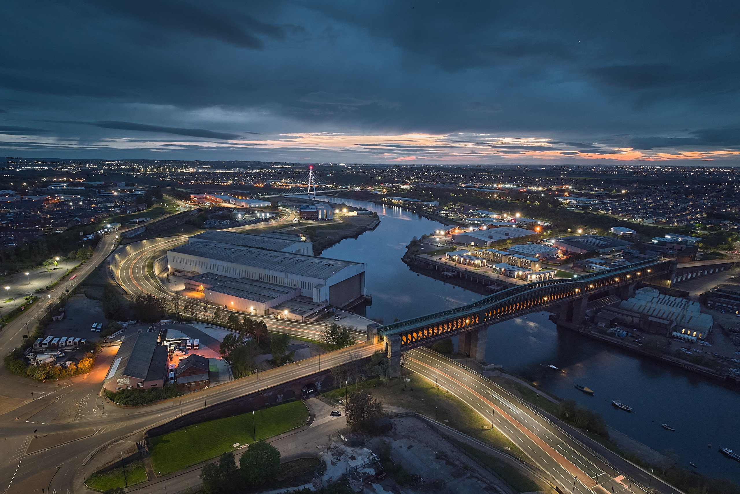 Aerial view of Queen Alexandra Bridge over the River Wear at dusk, city lights and traffic trails below, Northern Spire Bridge visible in the distance.