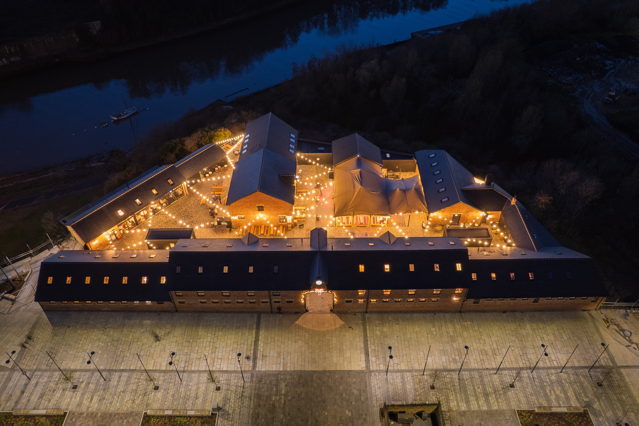 Aerial night view of the Sheepfolds warehouse courtyard strung with lights, beside the dark waters of the River Wear.